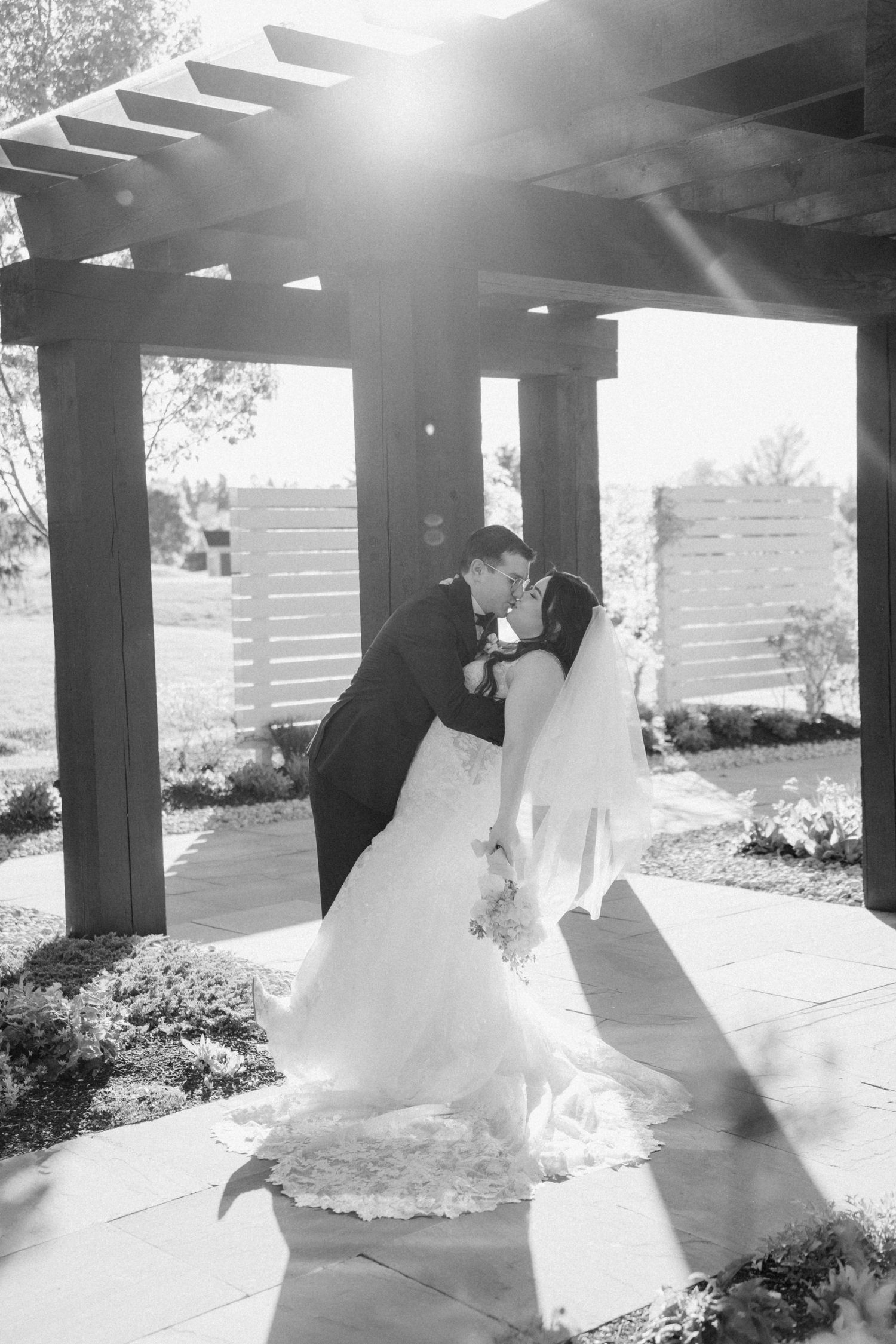 Romantic black and white photo of newlyweds sharing a kiss under a pergola with dramatic sunlight.
