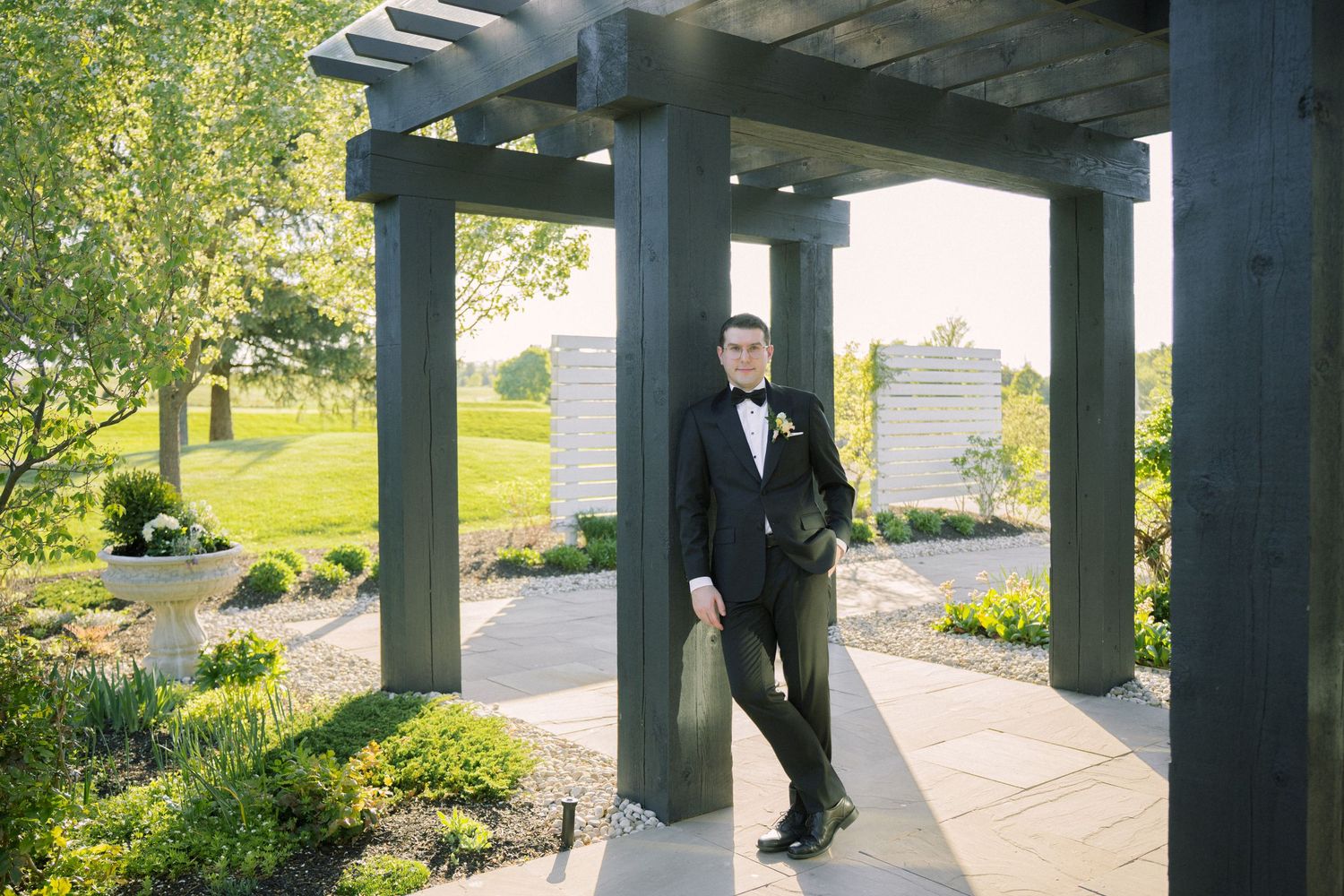 Formal portrait in a garden pergola with lush greenery and sunlit backdrop.