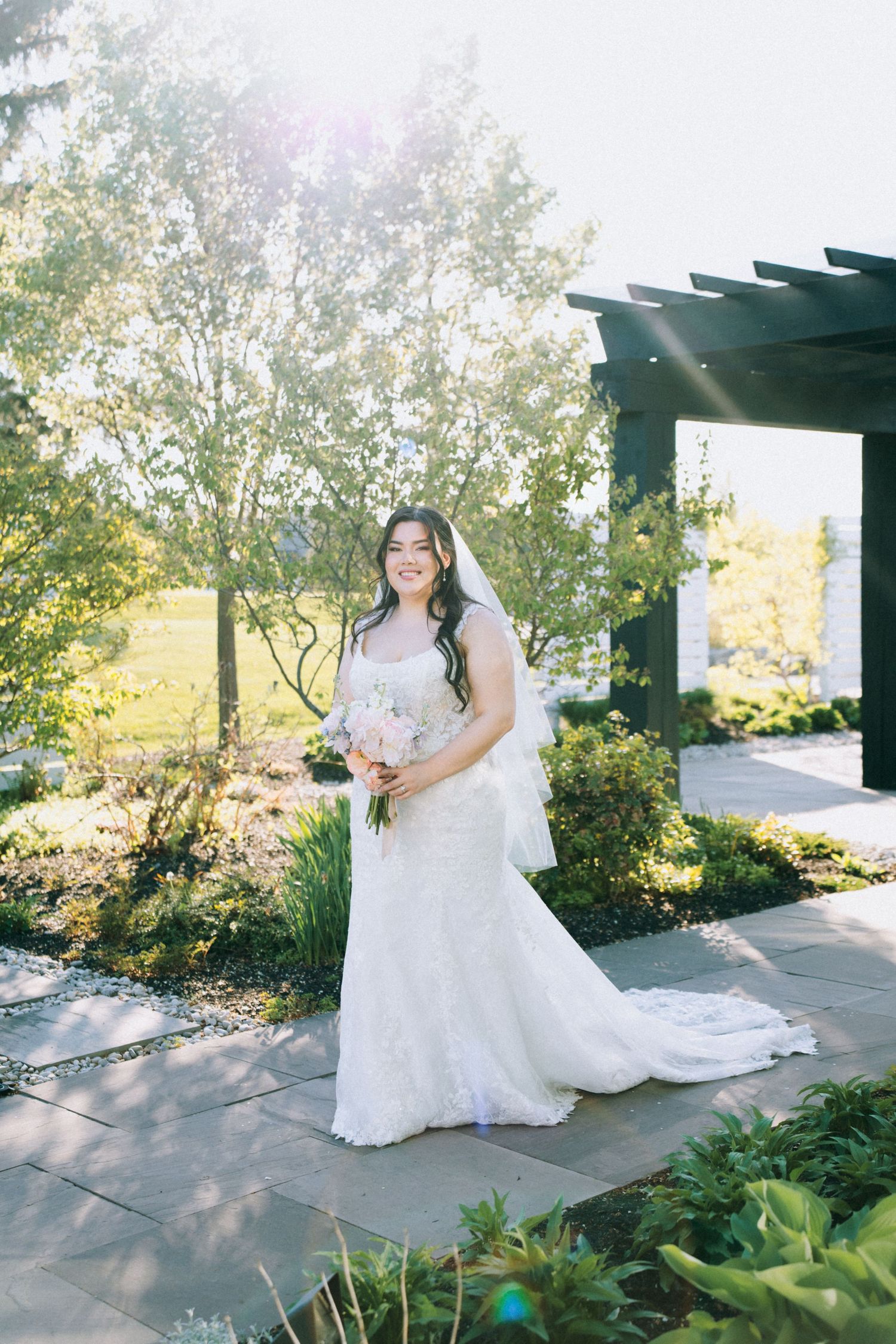 Elegant bride in a flowing white dress poses under a wooden pergola in a garden setting during a wedding photoshoot.
