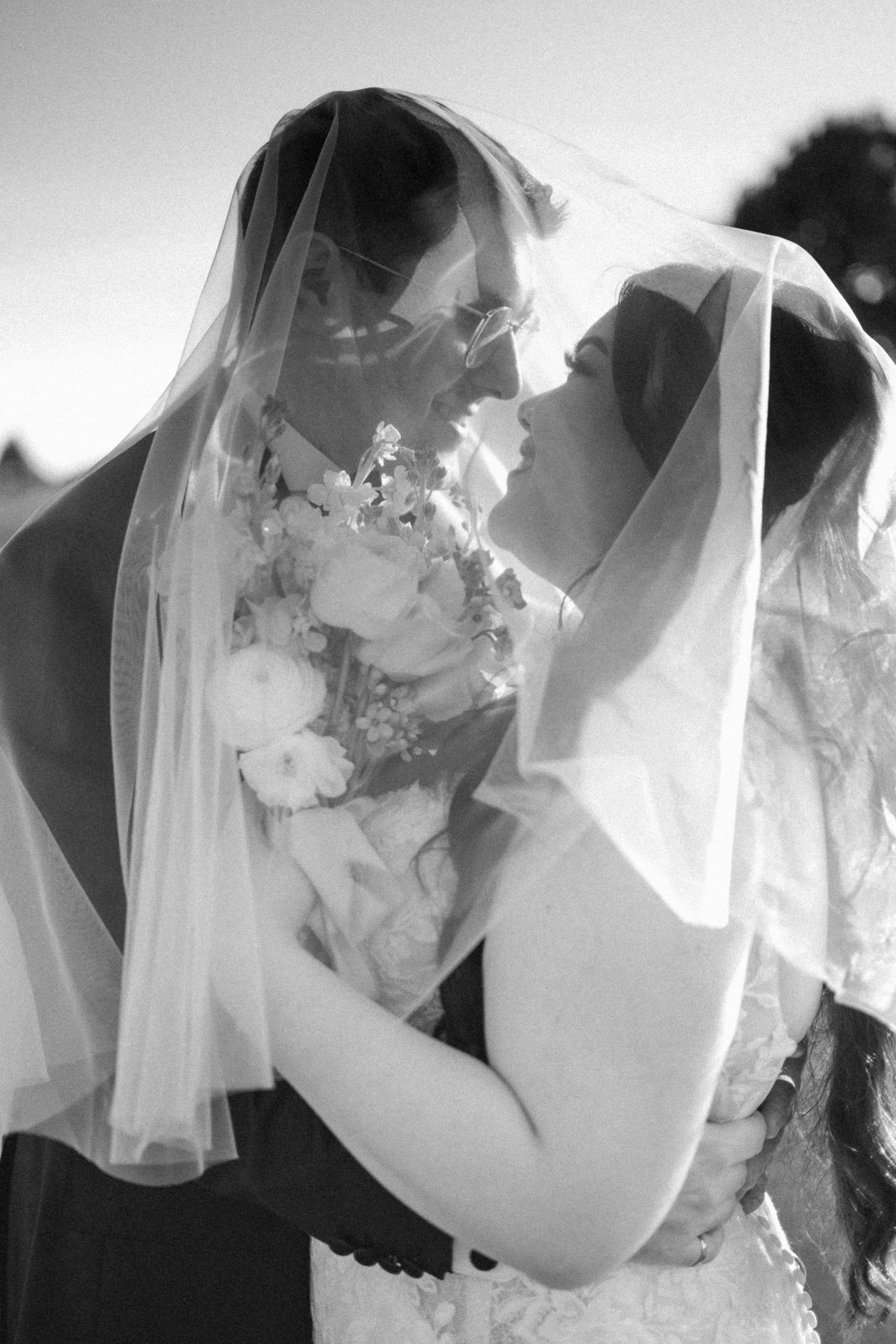 Intimate black and white portrait of couple behind bridal veil with floral bouquet details.