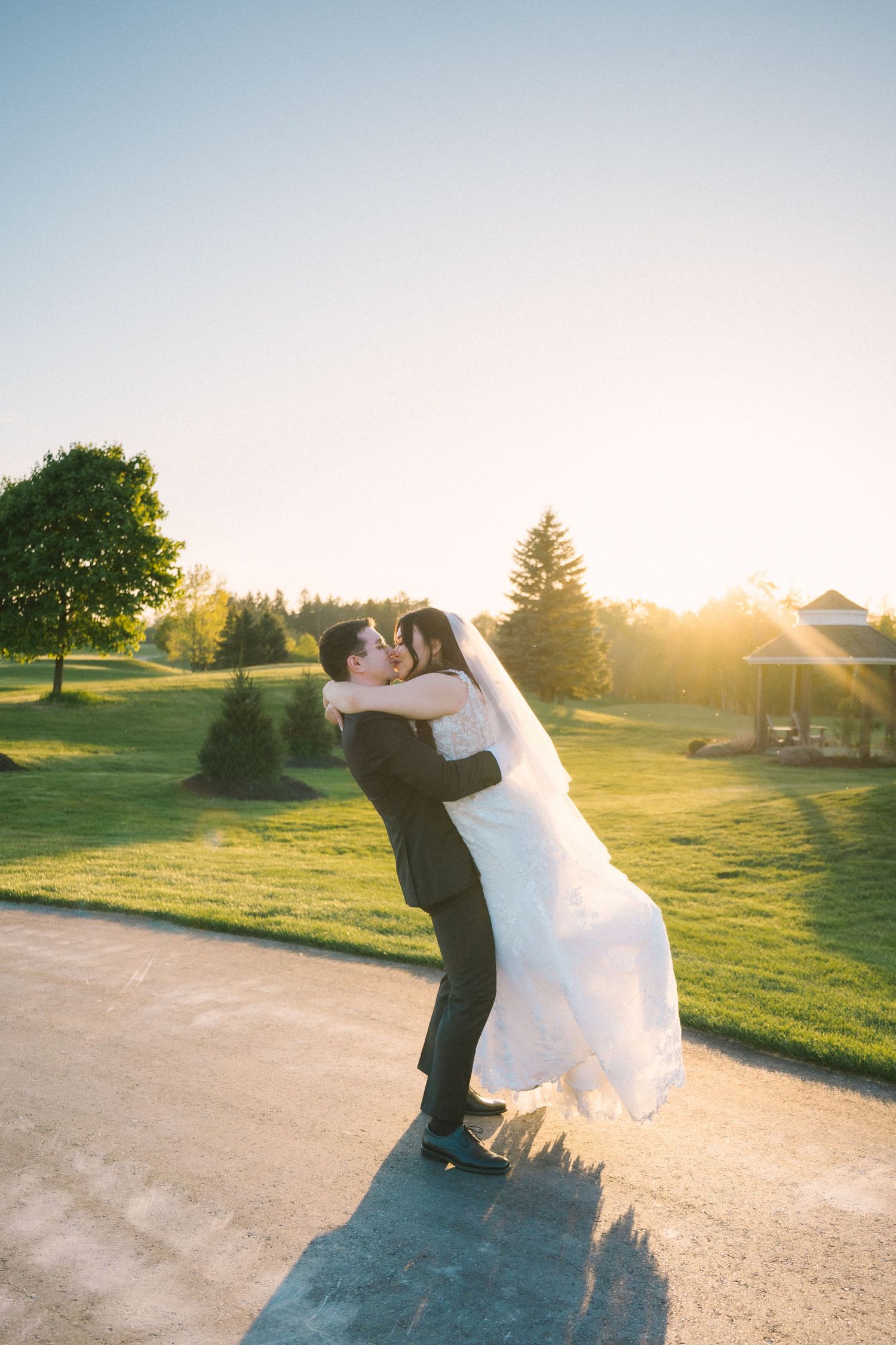 Sunset silhouette of newlyweds embracing on pathway with golden light and green landscape.