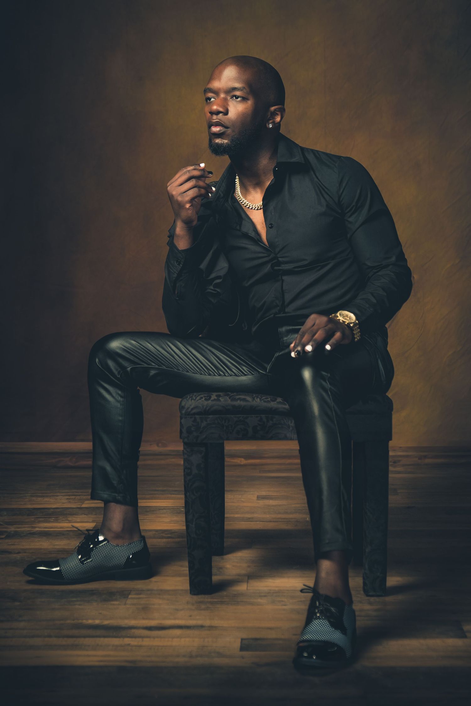 Stylish portrait of a person in black attire seated on wooden stool against dark backdrop with moody lighting.