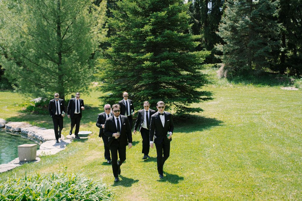 Groomsmen in black suits walking across a sunny green lawn.