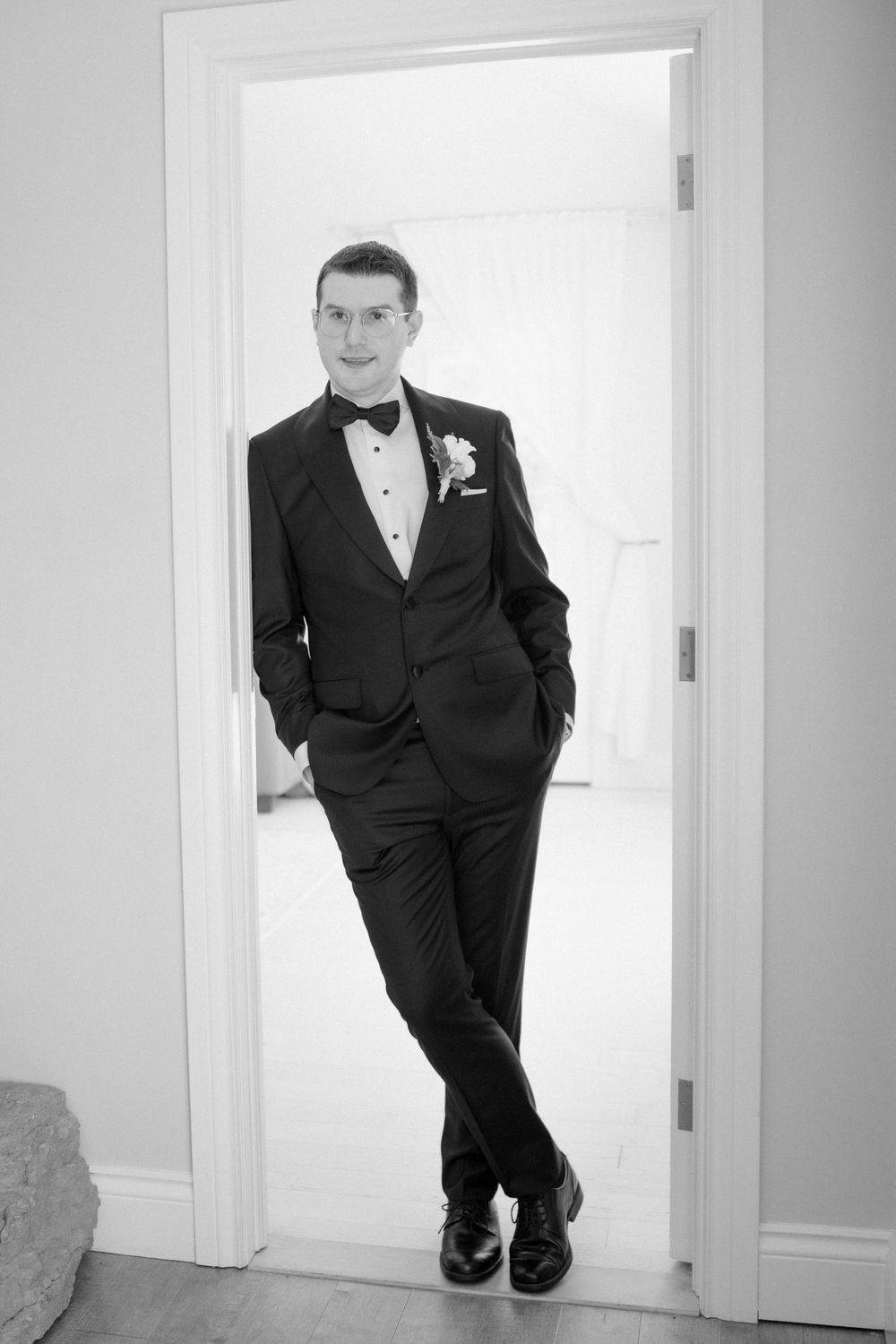 A groom in a classic black tuxedo and bowtie poses confidently in a doorway.