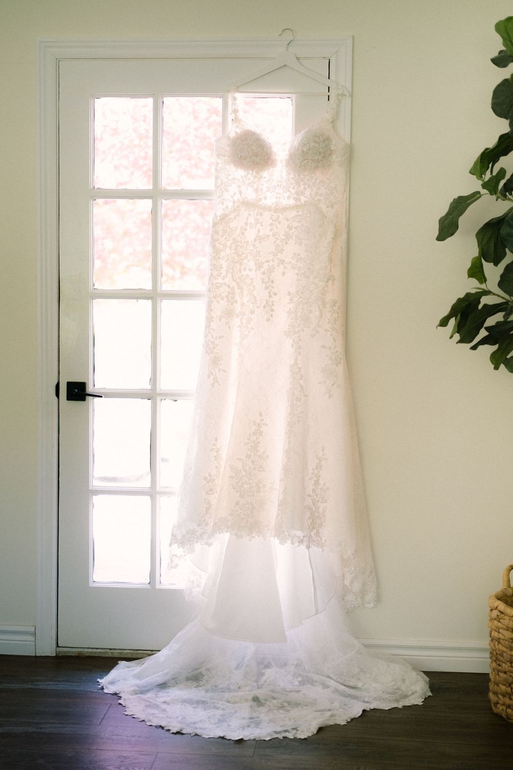 An elegant lace wedding dress hangs in front of a French door with natural light streaming through.