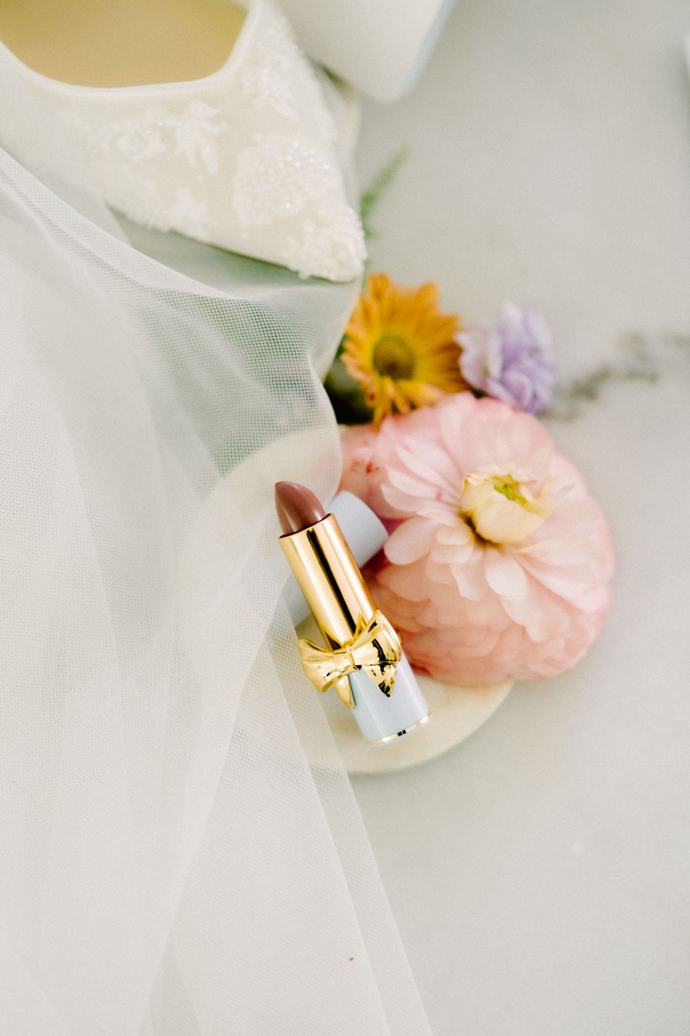 Wedding details with lipstick, delicate flowers, and tulle fabric styled in soft lighting.
