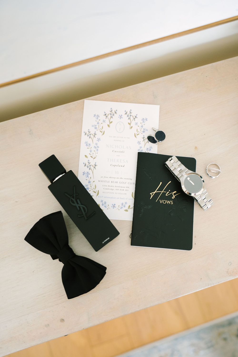 Wedding details including bow tie, invitation, vow books and watch on light surface.