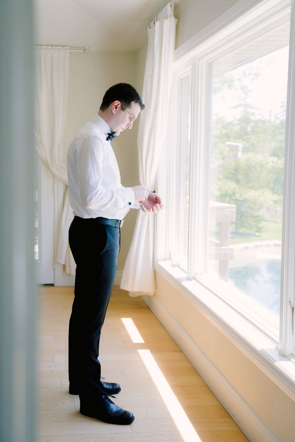 Person in formal attire standing by window adjusting cuffs.