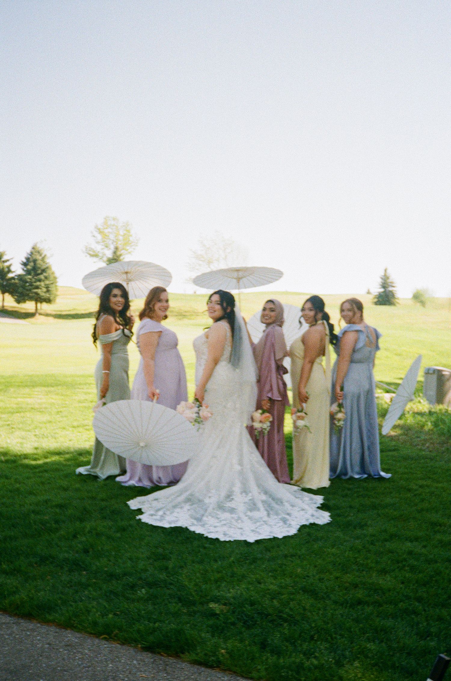 Bridesmaids in soft pastel dresses pose with parasols on green lawn.
