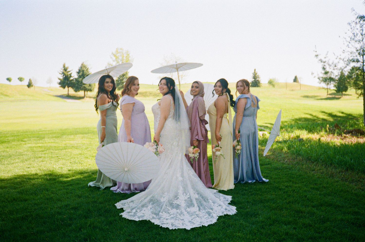 Wedding party poses on a lush green golf course with bridesmaids in pastel dresses surrounding a bride in a lace gown.