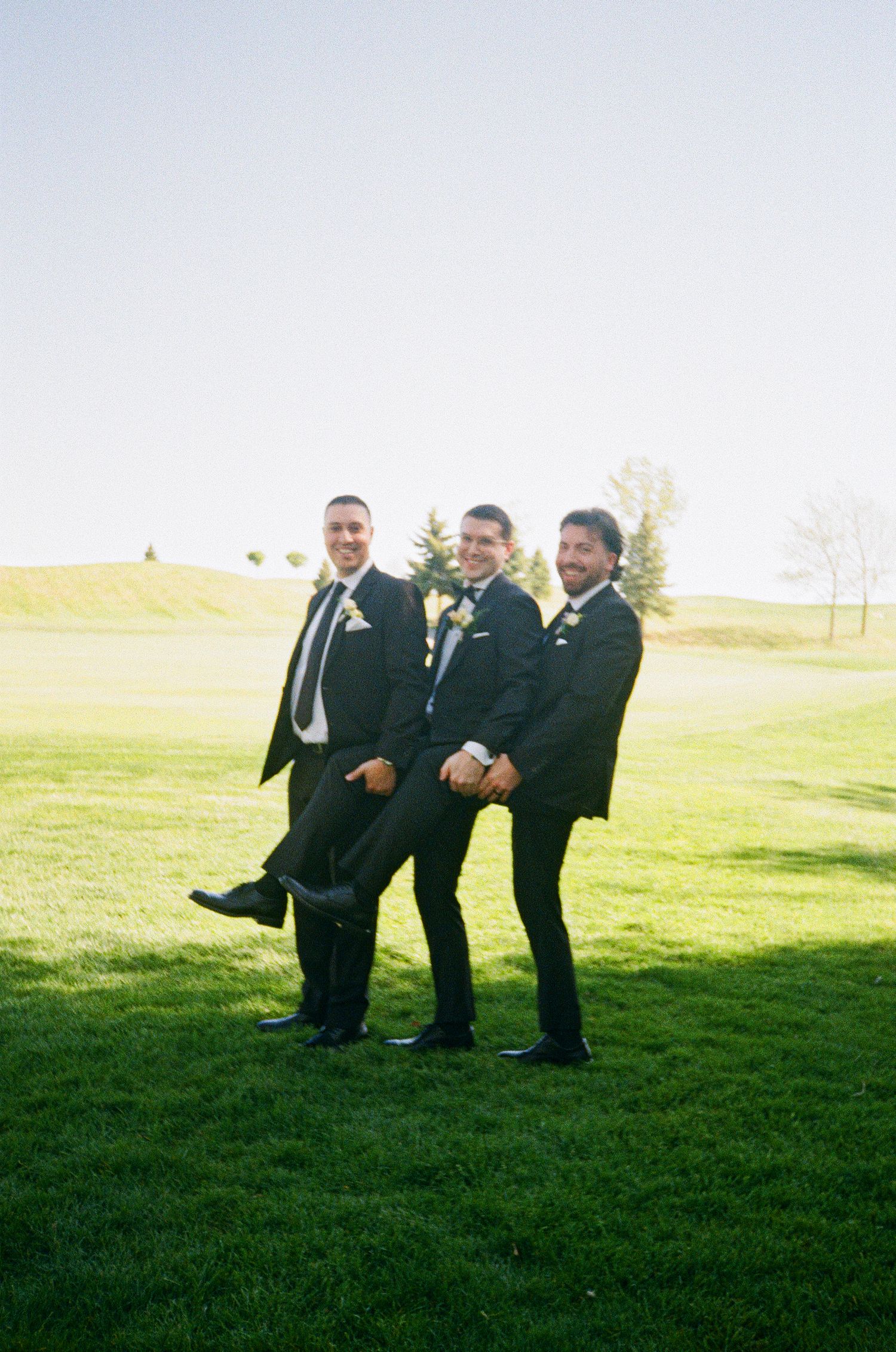 Three groomsmen in black suits strike a playful pose on a sunlit golf course.