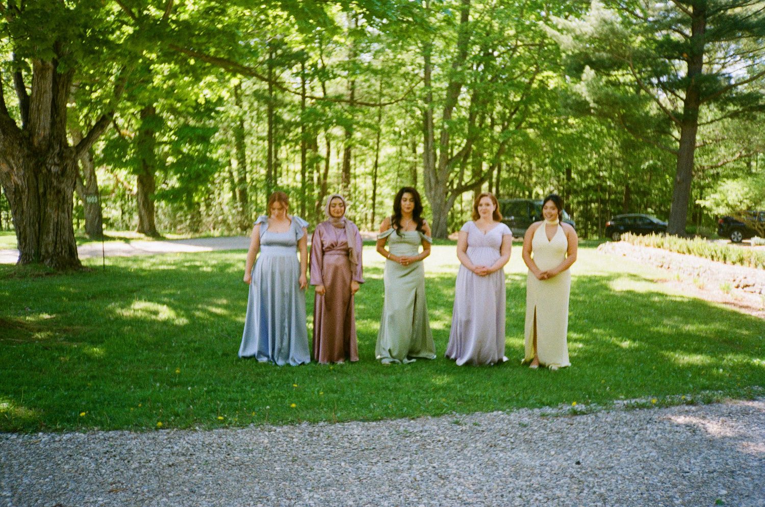 Bridal party in elegant pastel dresses poses together under green trees at an outdoor venue.