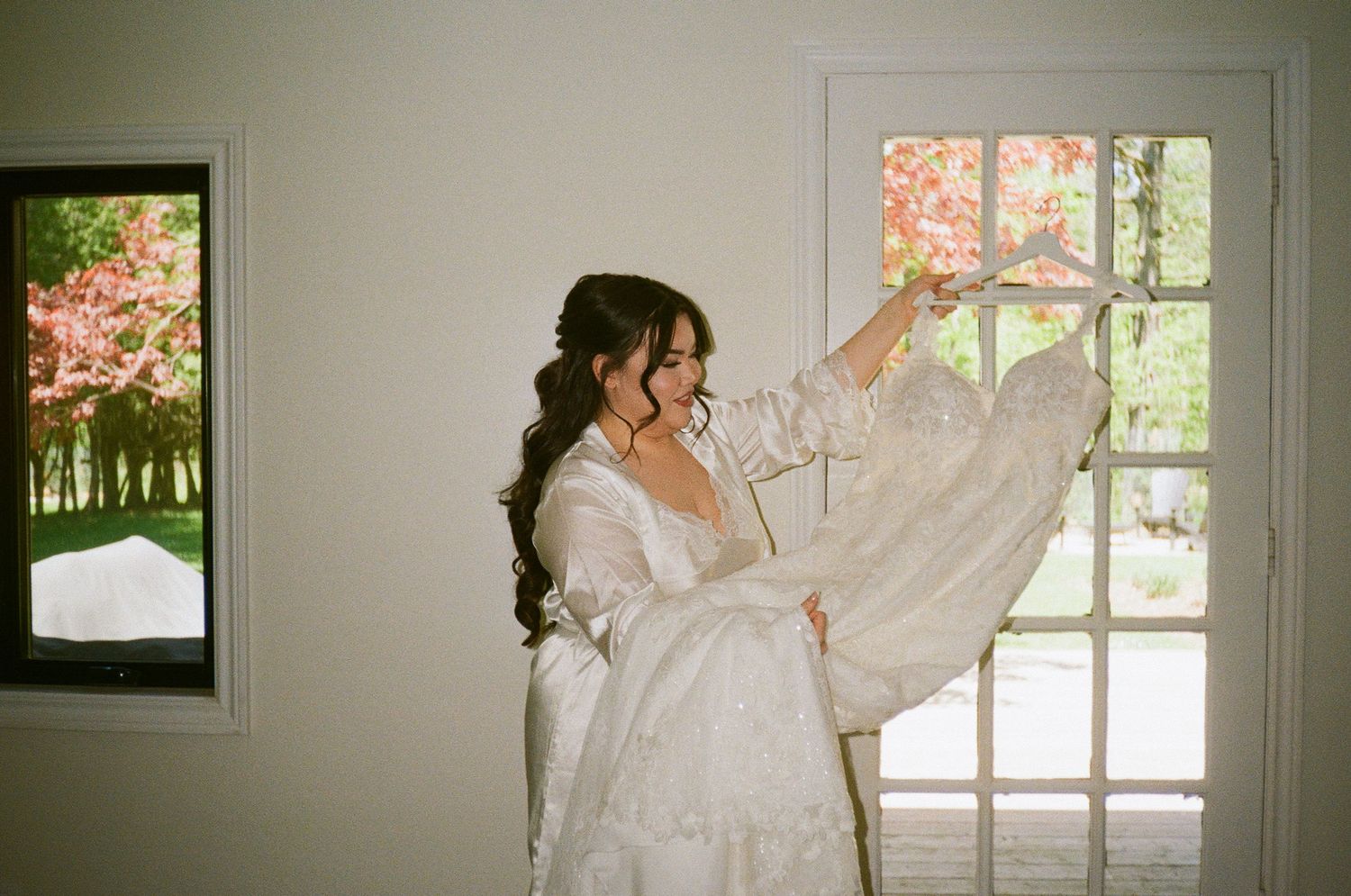 Getting ready scene by a bright window with white lace wedding dress on display.