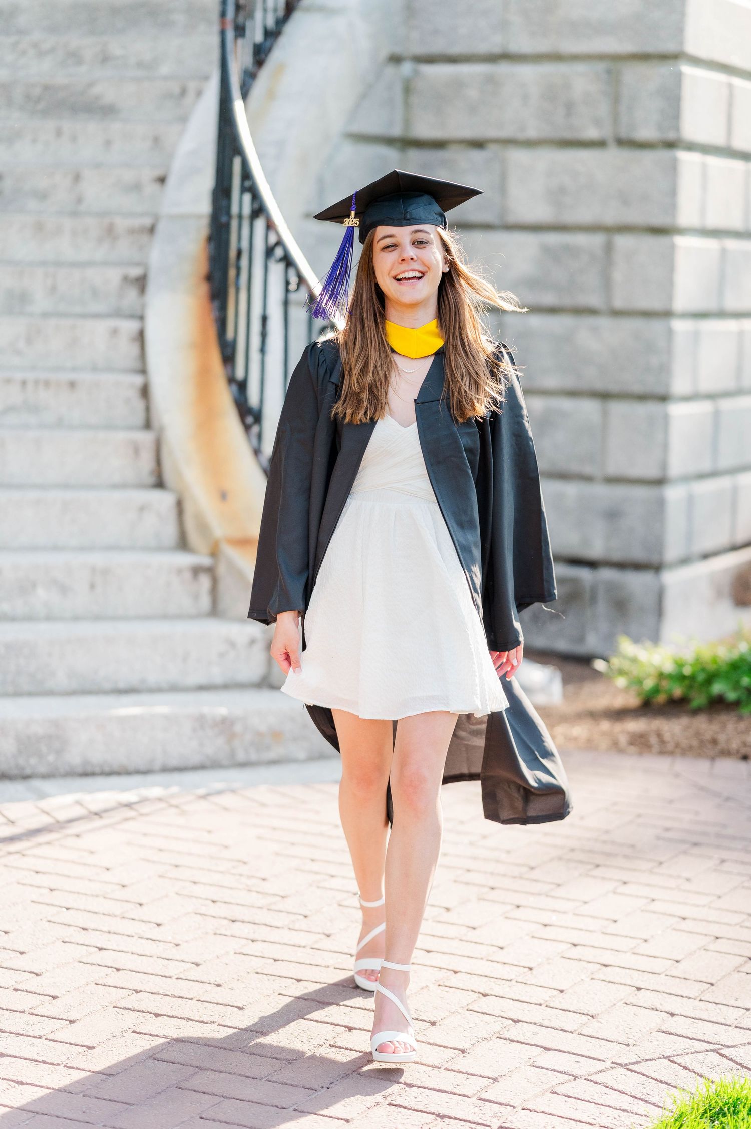 Graduate in cap and gown wearing yellow collar and white dress poses on brick walkway during commencement photo session.