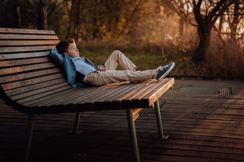 Persoon ontspannen op houten bank bij zonsondergang in herfstpark met blauwe jas en sneakers.
