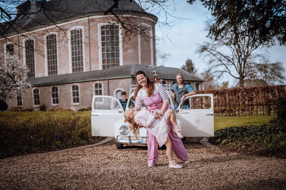 Een stel in roze outfits poseert romantisch bij een vintage witte auto voor een historisch herenhuis.