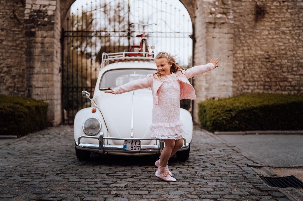 Een vrolijk persoon in een roze jurk danst naast een oude witte Volkswagen Kever die geparkeerd staat bij een stenen poort.