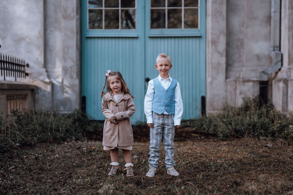 Twee jonge kinderen in stijlvolle casual outfits staan naast elkaar voor een vintage turquoise deur en stenen muur.