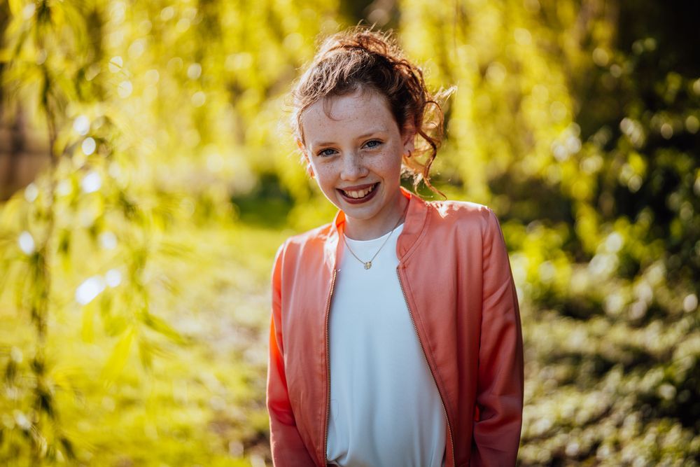 Een jong individu met een koraalkleurig vest en wit overhemd lacht stralend tegen een zonovergoten gele achtergrond van wilgenbomen.