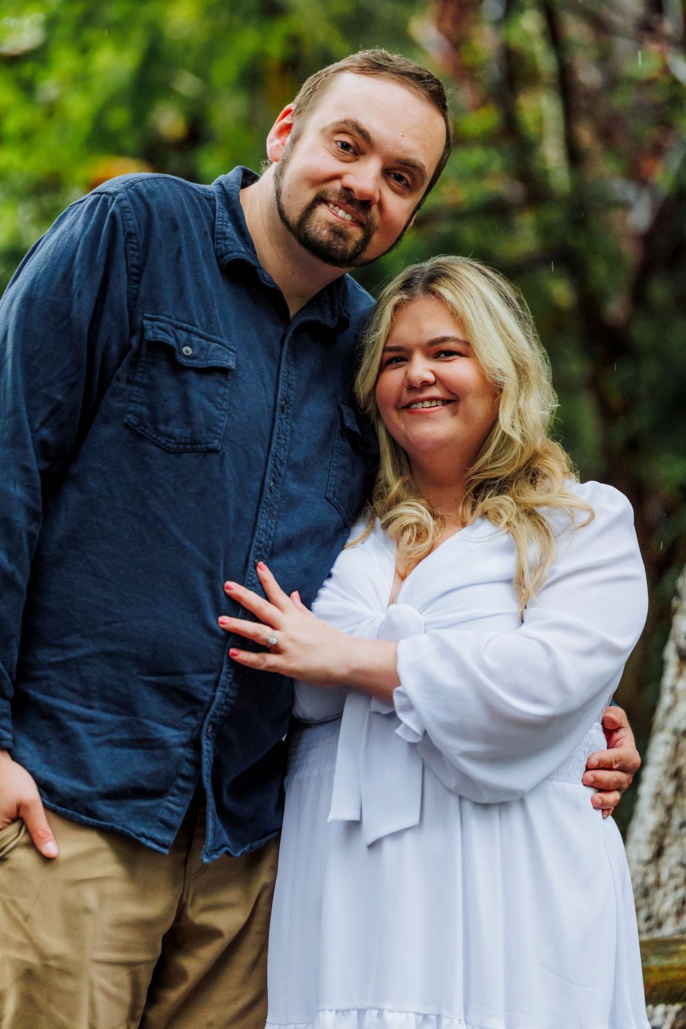 A couple poses together outdoors wearing a navy blue shirt and white dress against a green natural background.