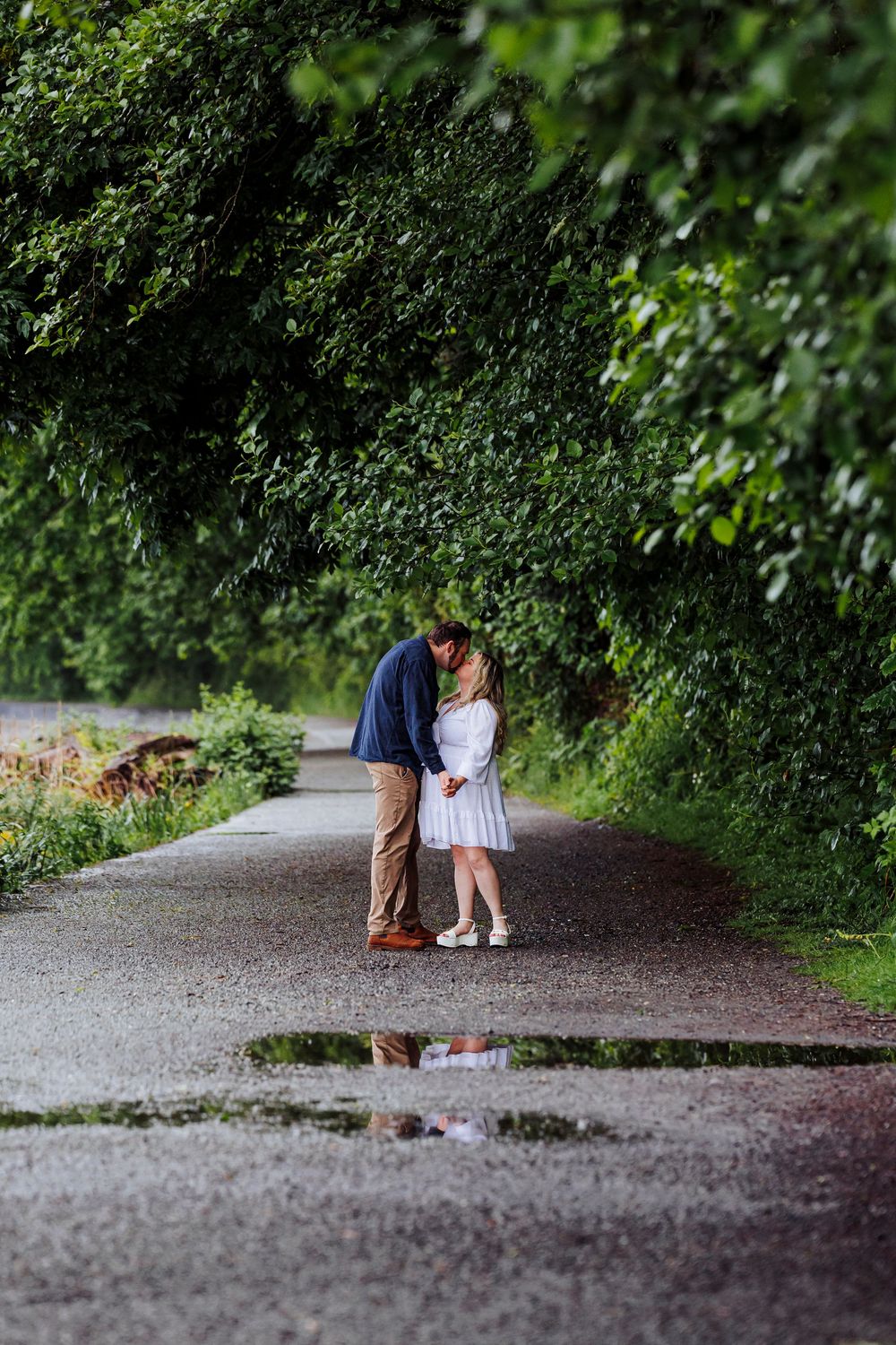 A couple embraces on a wet path surrounded by lush green foliage and trees in a romantic garden setting.