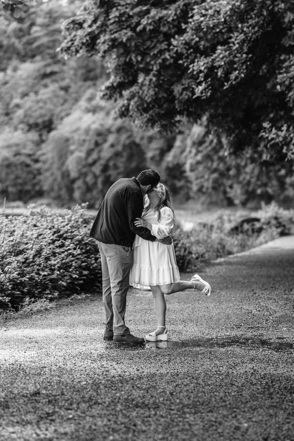 A romantic black and white moment captured on a tree-lined path as two figures embrace in an intimate outdoor setting.