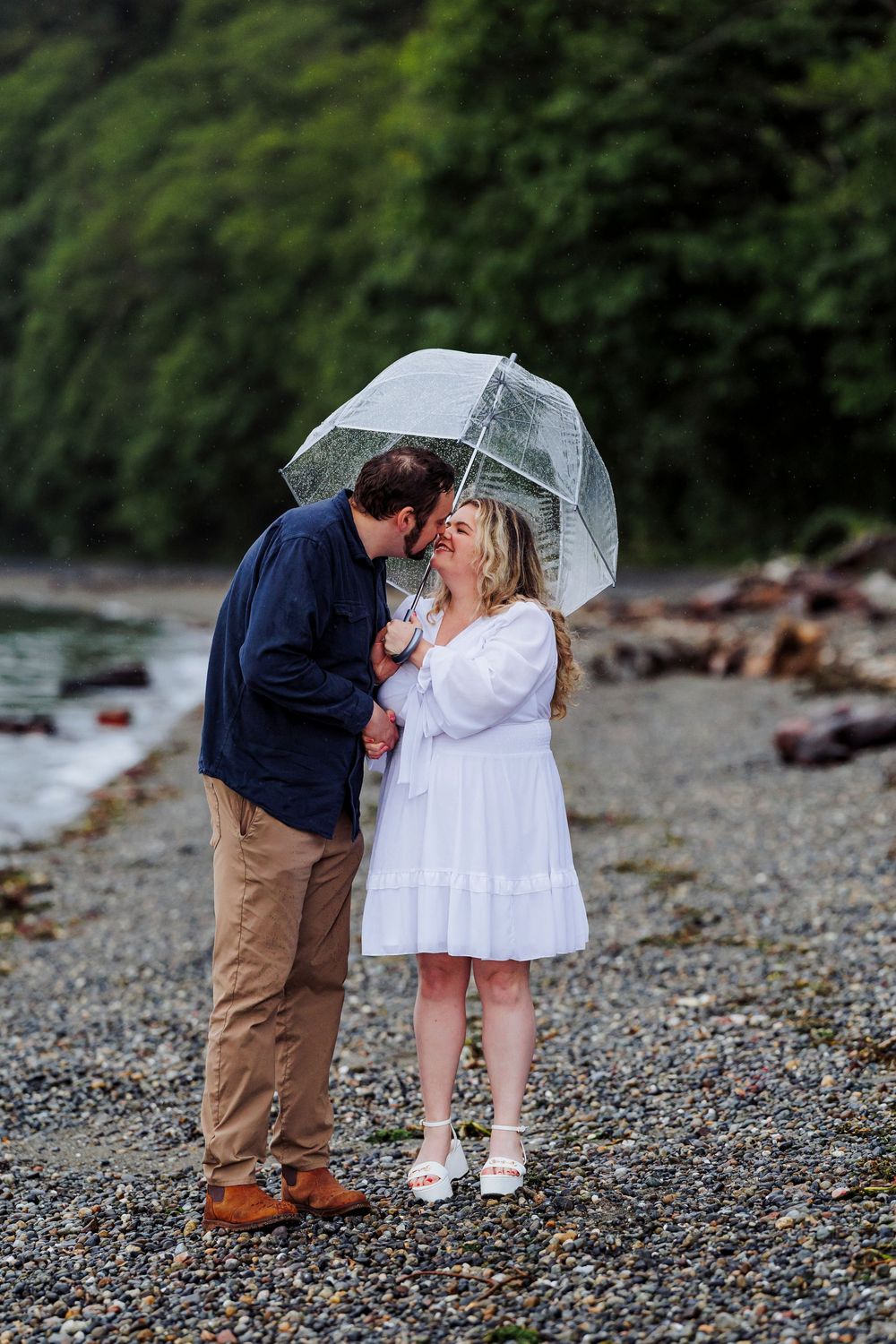 Romantic couple sharing an intimate moment under a clear umbrella on a rocky beach.