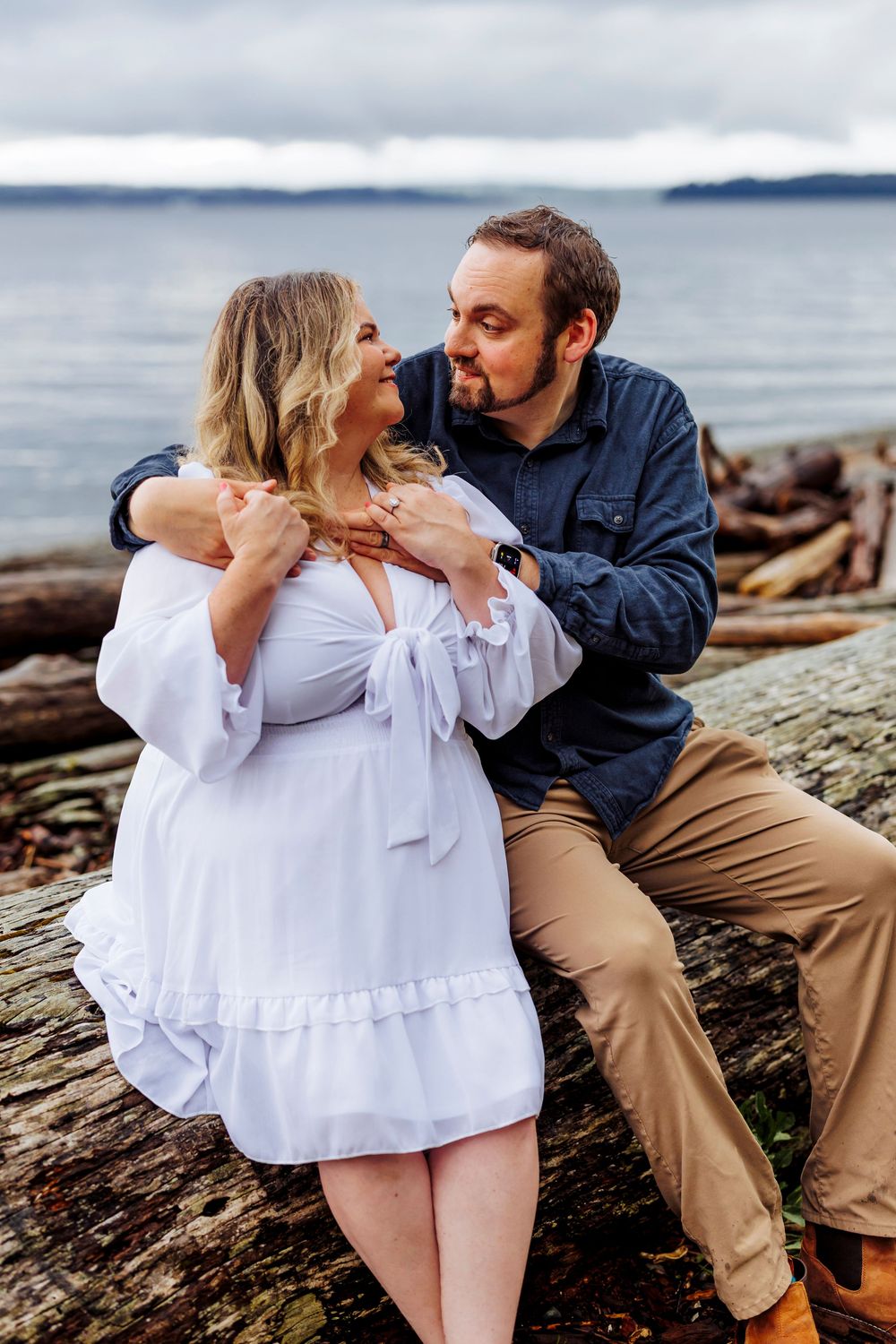 A romantic couple embraces on driftwood by the ocean during a cloudy day photoshoot.