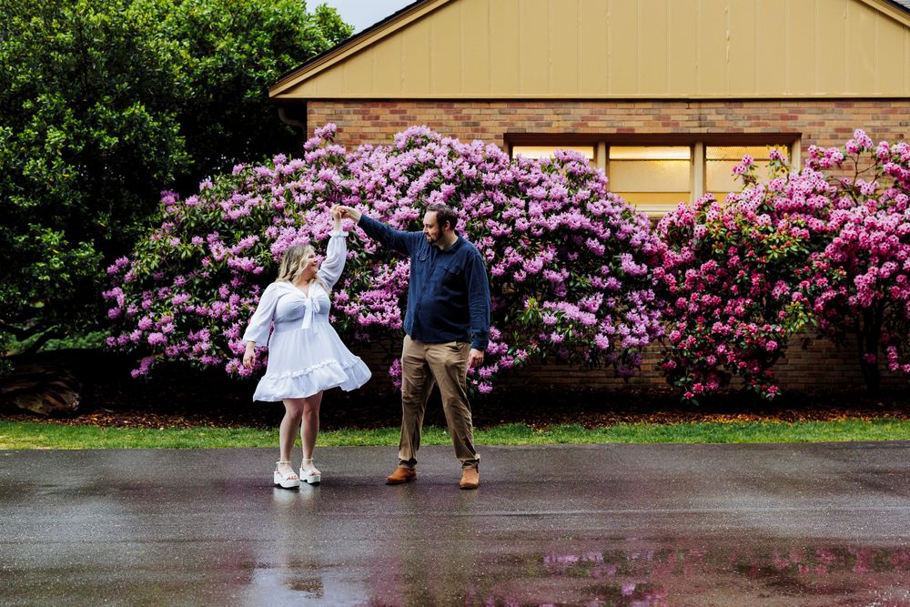 A couple dances in front of vibrant purple rhododendron bushes blooming outside a house on a rainy day.