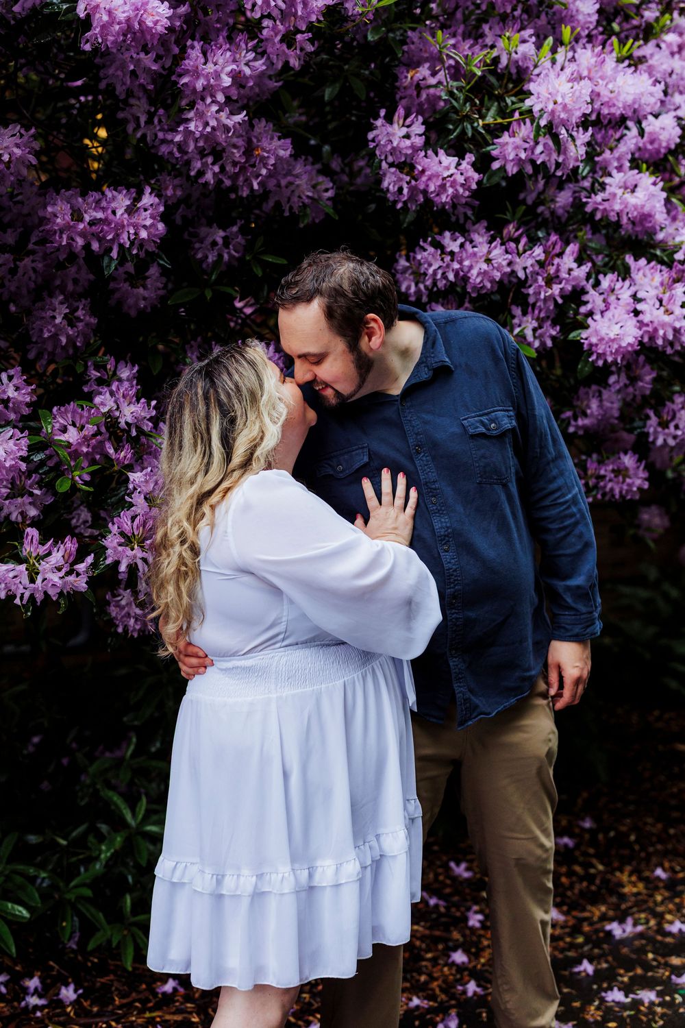 A couple shares an intimate moment in front of blooming purple rhododendrons in a spring garden.