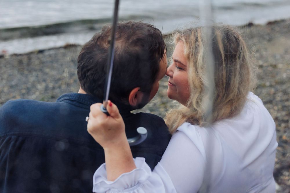 Couple sharing romantic moment on beach with an umbrella, woman in white and partner in navy clothing.