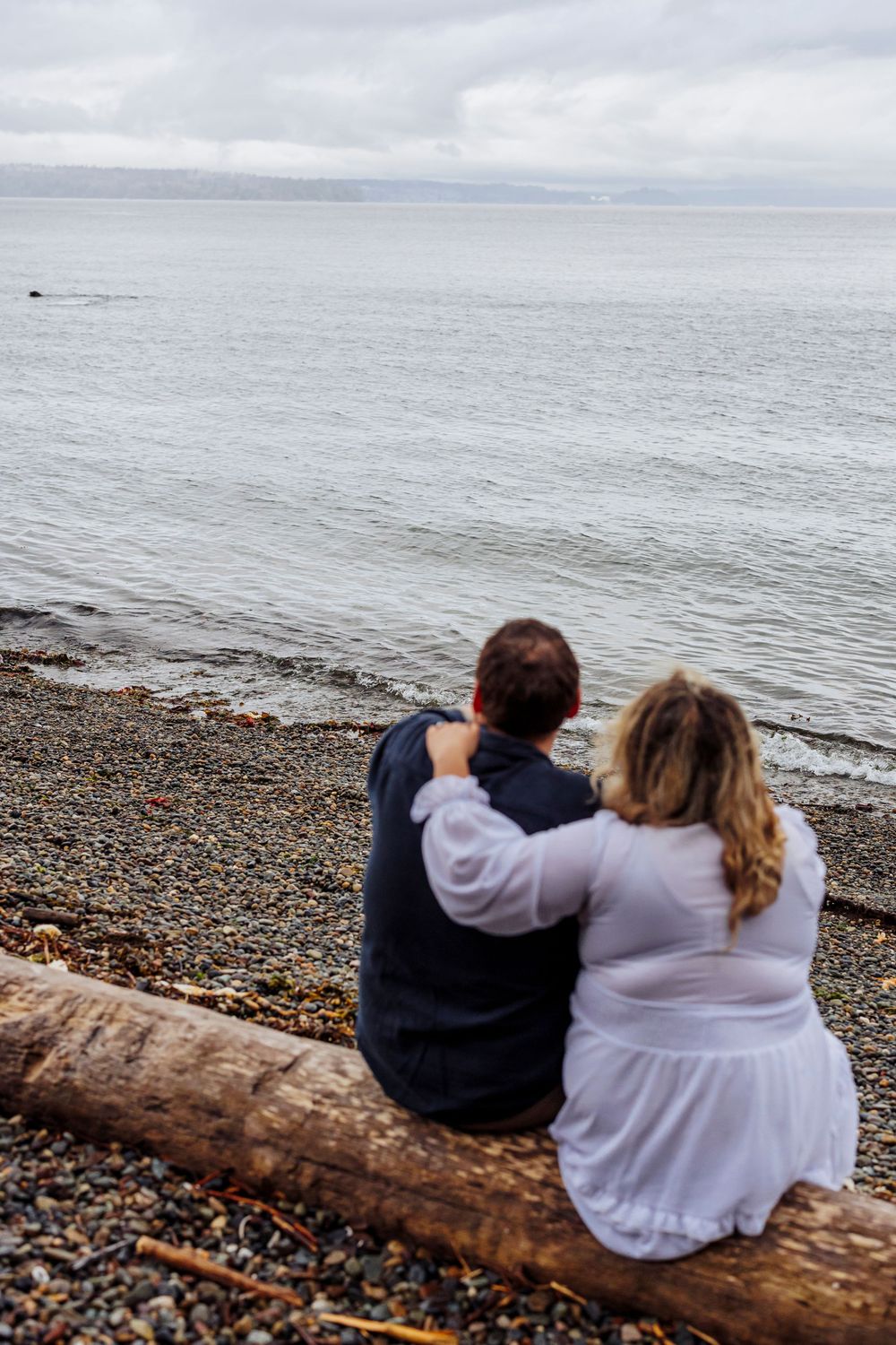 A couple sits together on a log at a rocky beach overlooking the ocean on an overcast day.