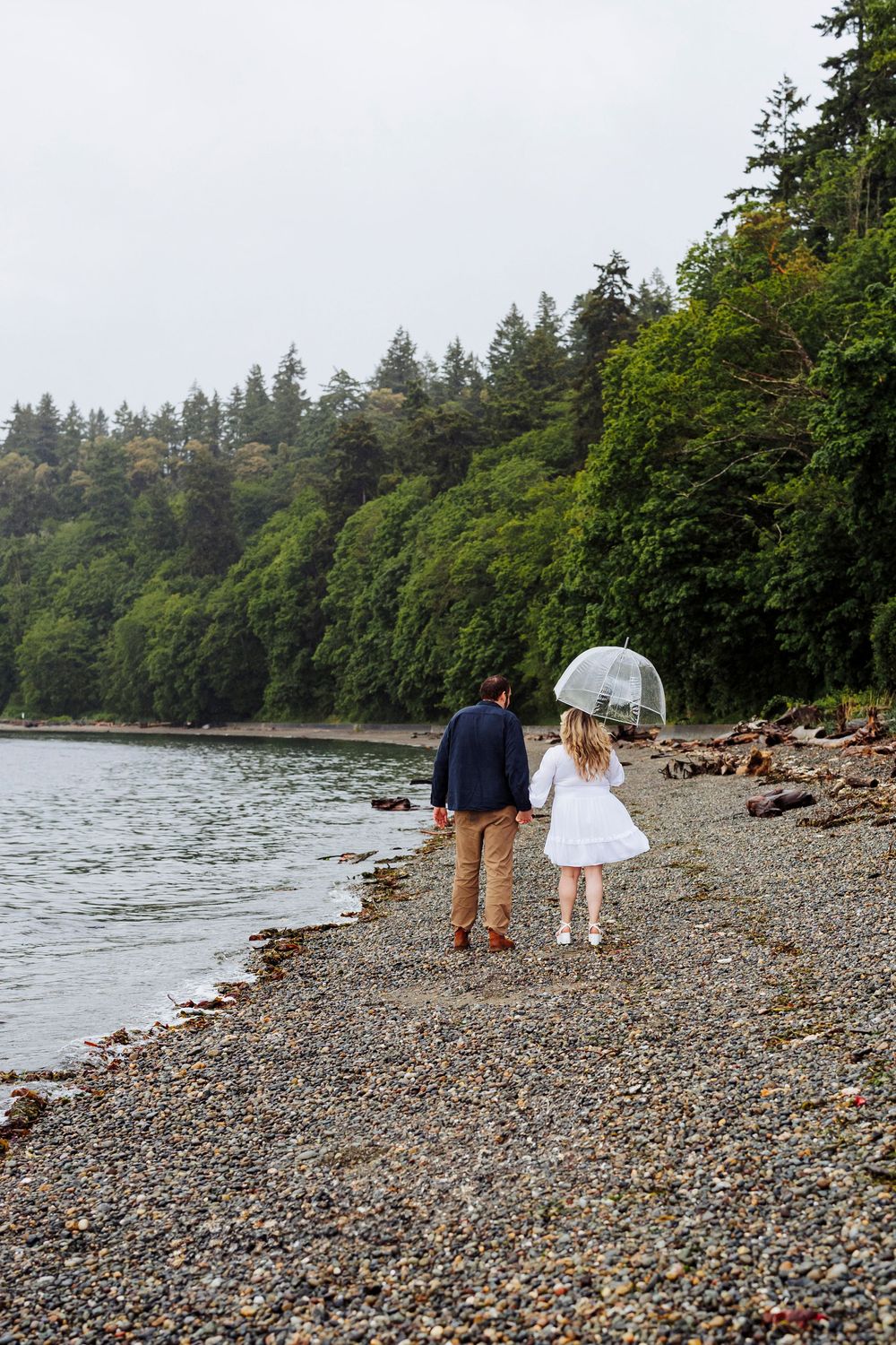 Couple walking together along a rocky beach shoreline surrounded by evergreen trees on a cloudy Pacific Northwest day.