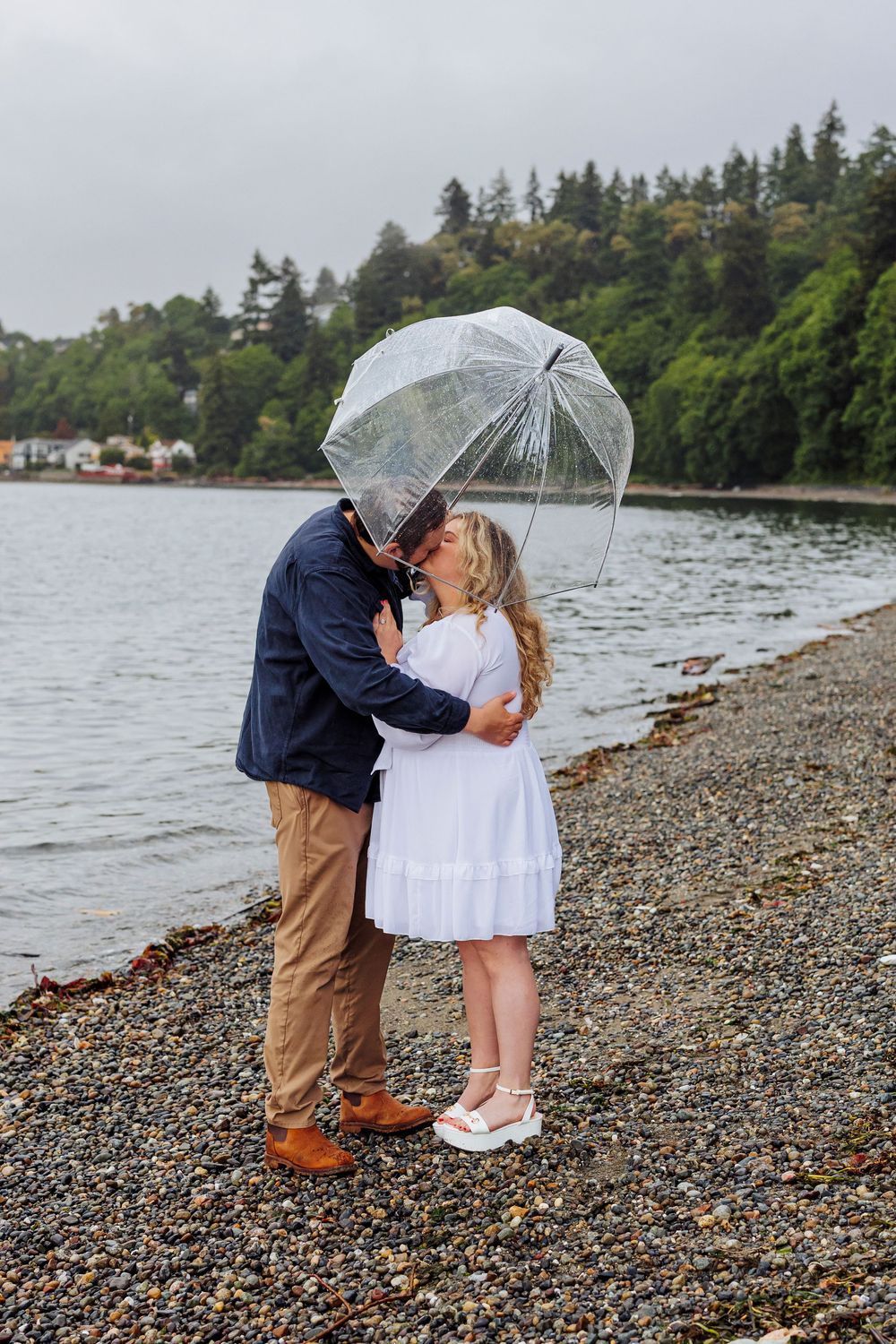 A couple shares a romantic kiss under a clear umbrella on a rocky beach shore with evergreen trees in the background.