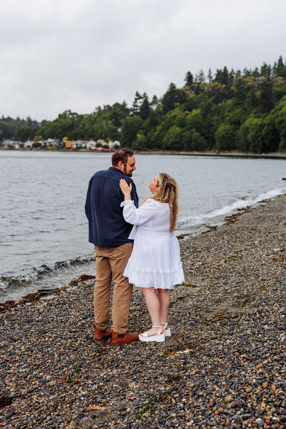 A couple embraces while standing on a rocky beach shore with evergreen trees and boats visible in the background.