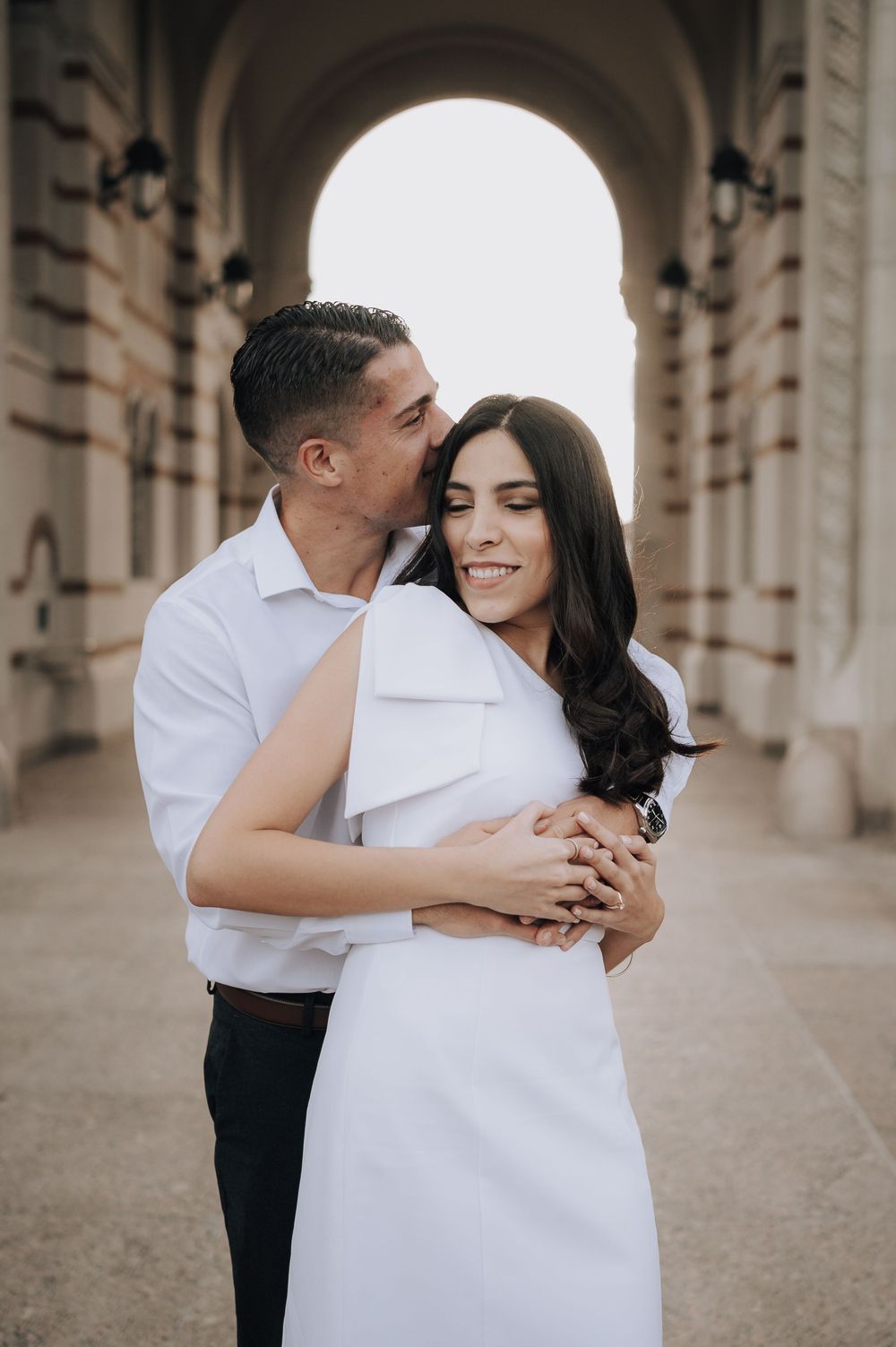 Romantic couple in white outfits embrace under an ornate stone archway with soft lighting creating an intimate moment.