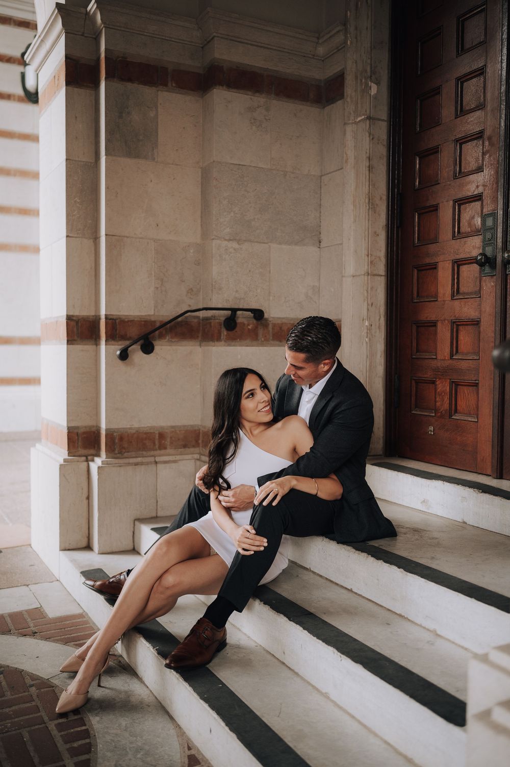 A romantic couple embraces on stone steps outside an elegant building with a wooden door.