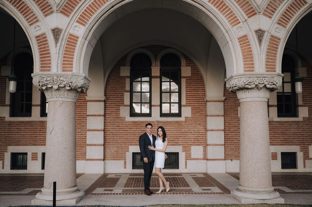 A couple stands between ornate stone columns beneath an arched brick facade with decorative windows at a university building.