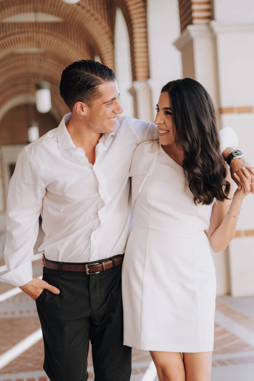 Couple in coordinating white outfits sharing a romantic moment while walking through an arched corridor.