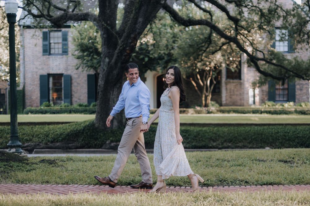 A couple strolls together across a green lawn in front of a historic pink stucco building with overhanging oak trees.