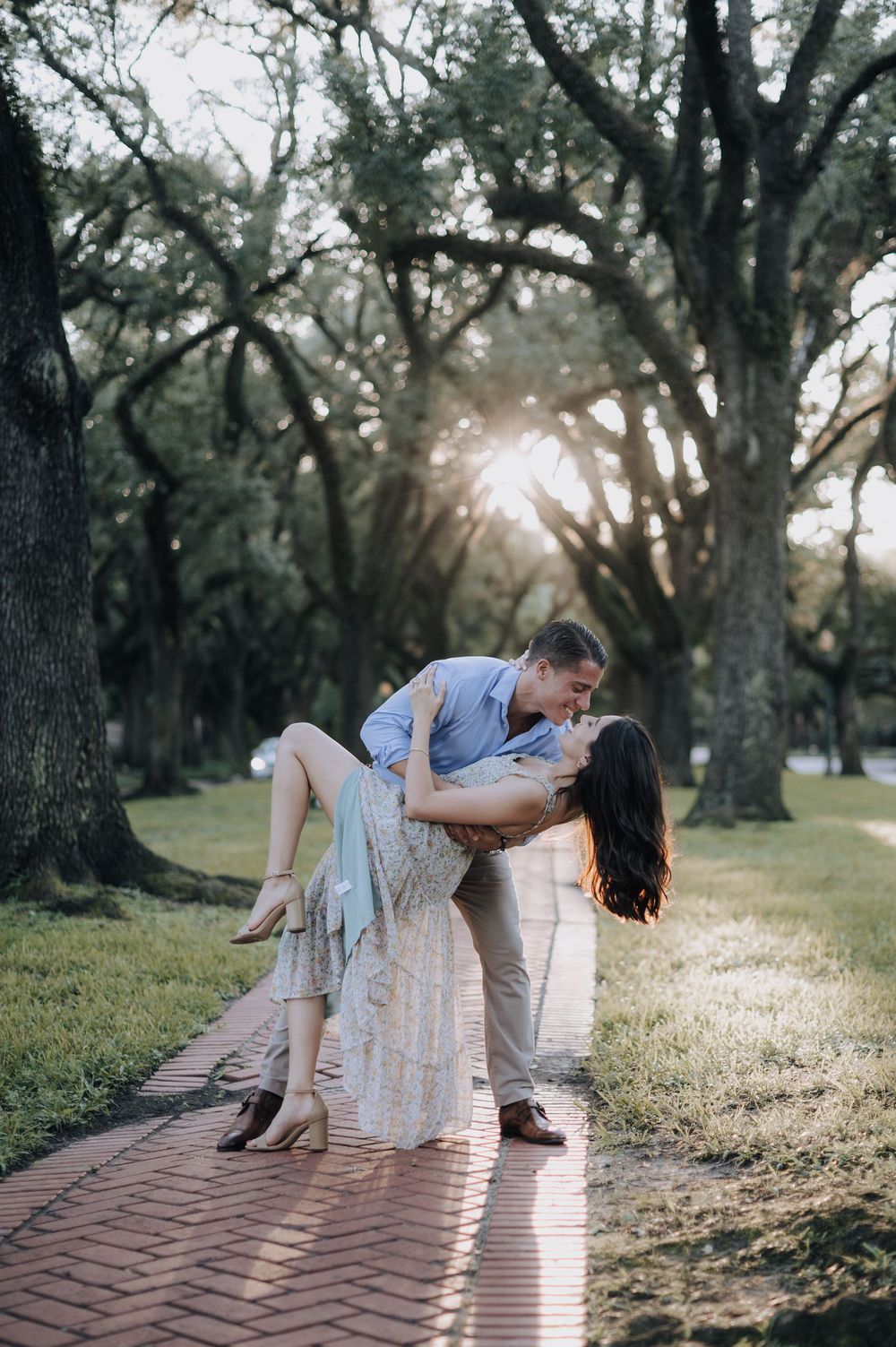 A romantic couple shares an intimate dip pose on a sunlit brick pathway surrounded by oak trees in a park.