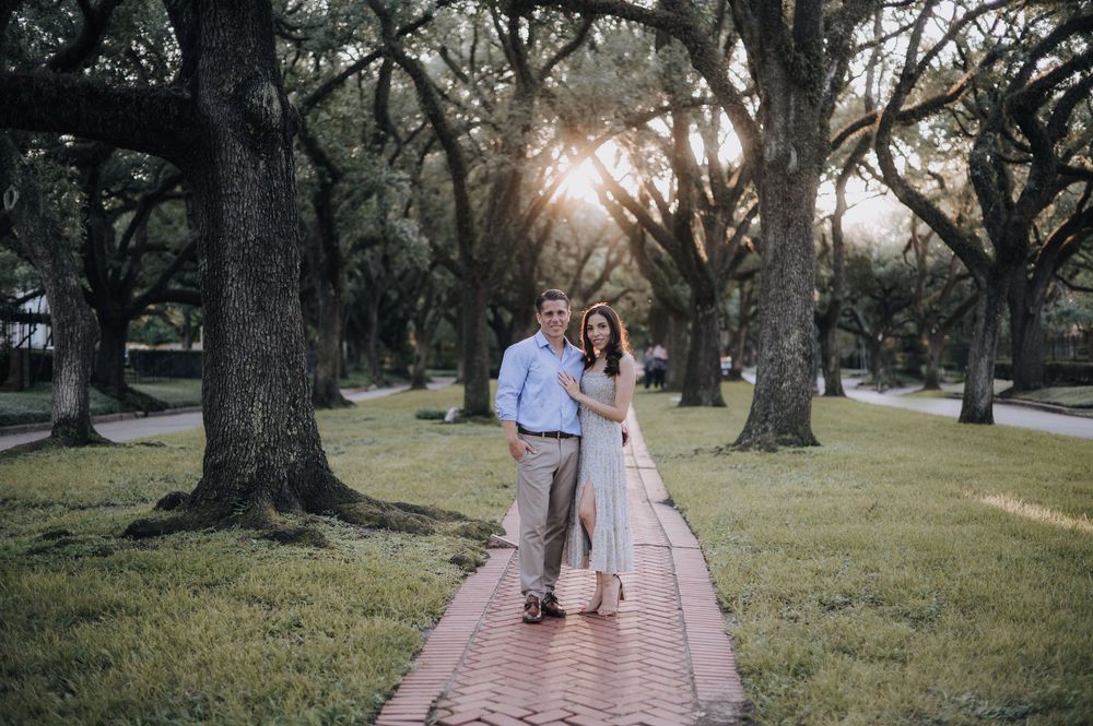 A couple walks along a brick pathway through a park with sunlight streaming through moss-draped oak trees.