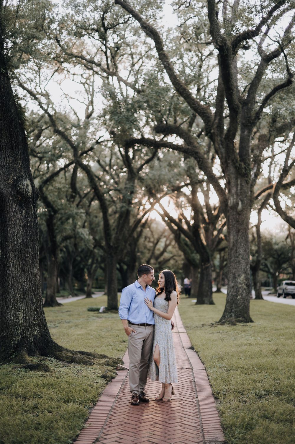A couple walks together on a brick path surrounded by towering oak trees in a peaceful park setting at dusk.