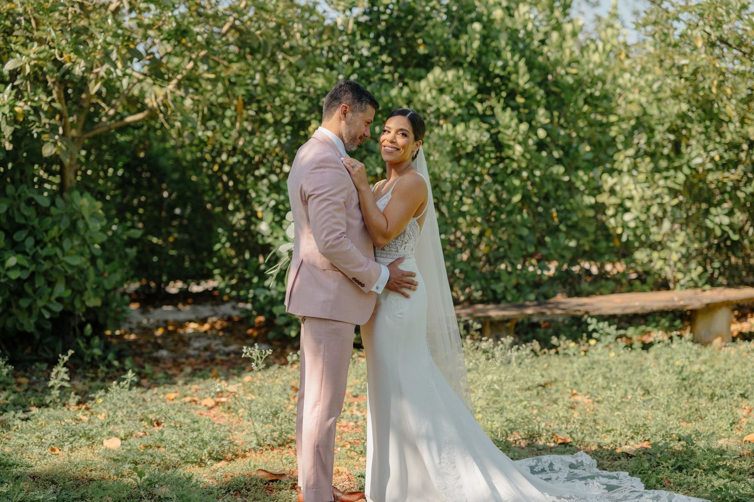 A couple enjoys an intimate moment on their wedding day in a sunlit orchard garden setting.
