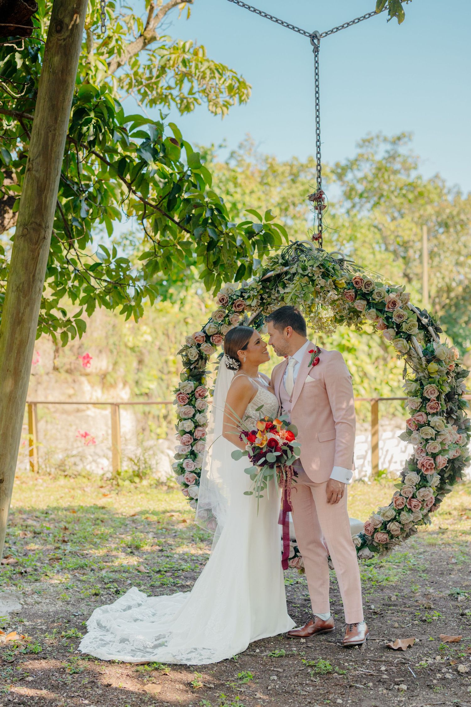 Wedding couple sharing a romantic moment under a floral arch decorated with pink roses in a garden setting.