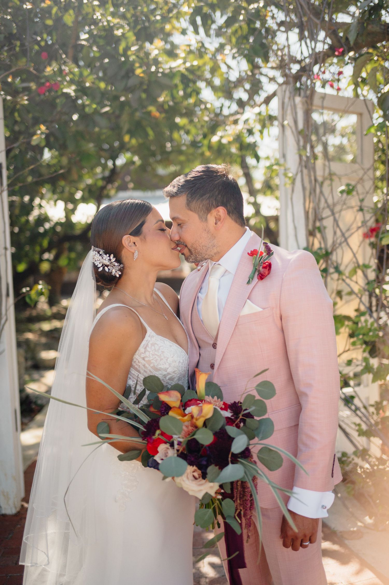 Sun-lit garden wedding scene with couple in white dress and blush pink suit sharing tender moment with colorful bouquet.