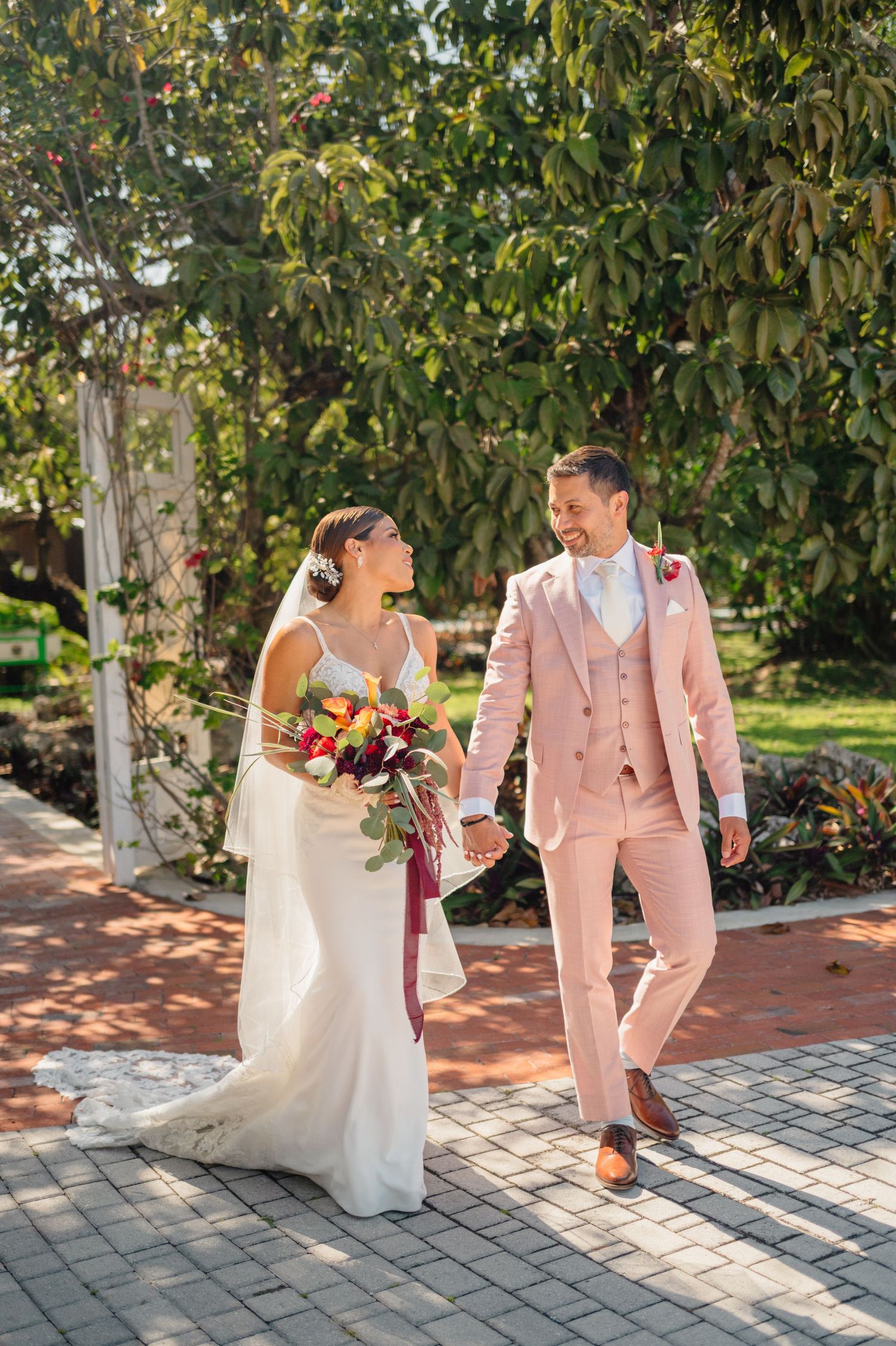 A couple in wedding attire strolls on a paved path, with the groom wearing a pink suit and bride holding a colorful bouquet.