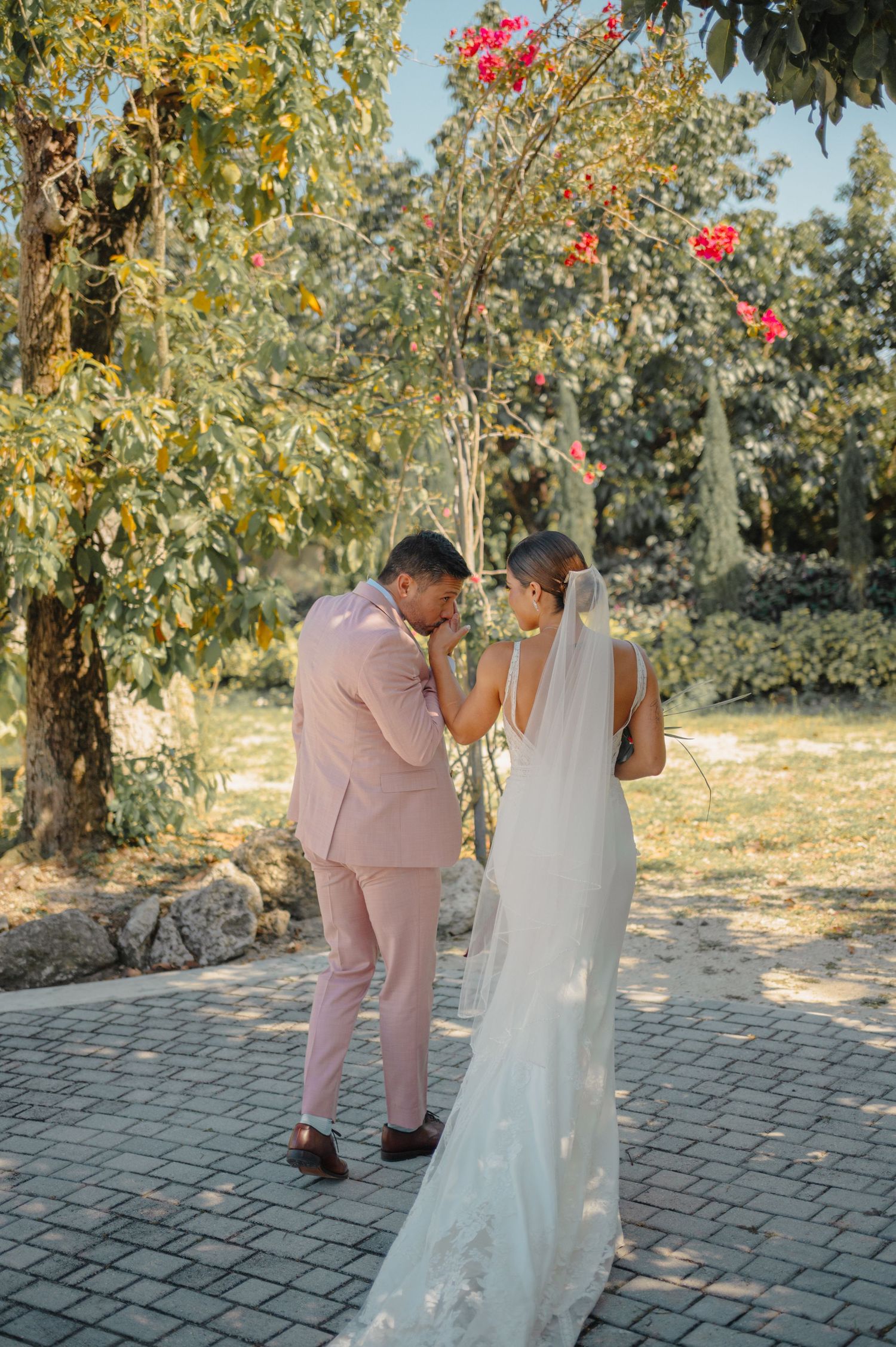 A romantic garden wedding scene with a couple in pink suit and white dress on a brick path surrounded by blooming trees.