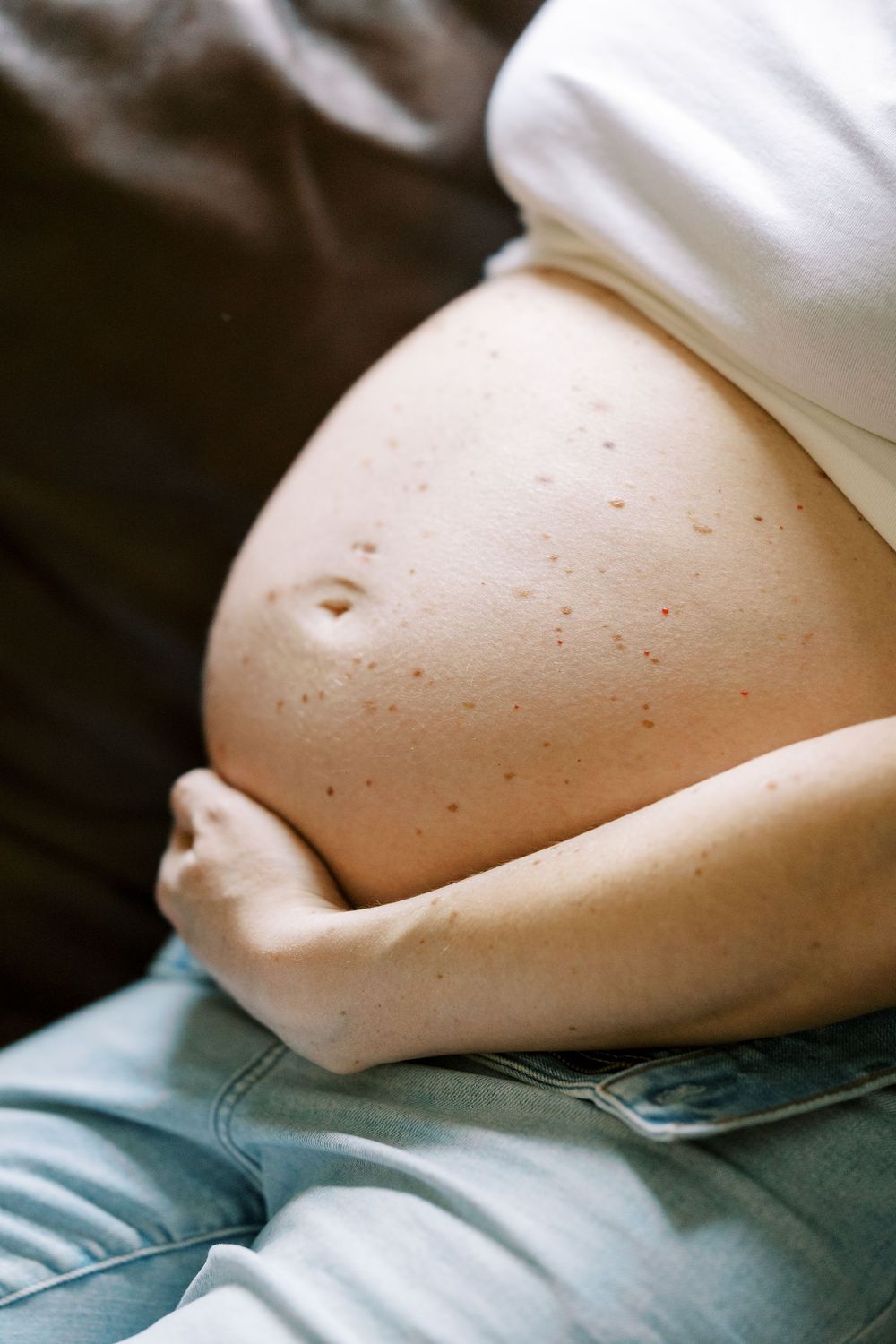 Close up of pregnant belly with woman wearing white shirt and jeans while lounging on dark fabric in Clifton, New Jersey.