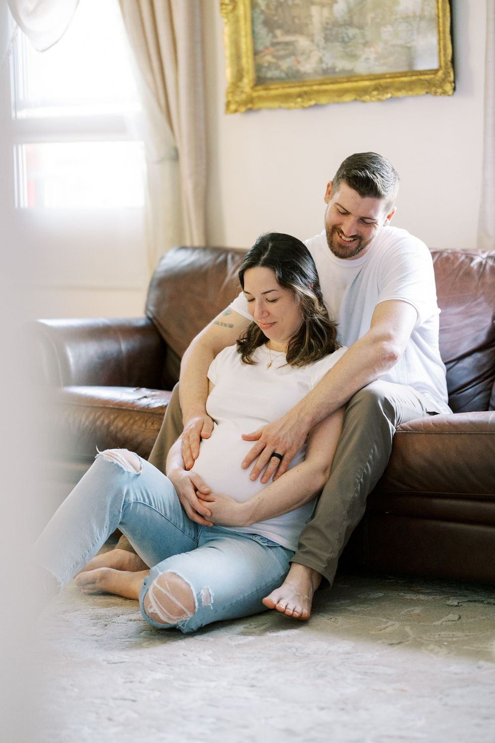 Expecting couple relaxes on leather couch as they lovingly embrace the pregnant belly in a sunlit living room in Clifton, New Jersey.